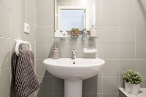 Une salle de bain avec un lavabo blanc, un miroir au-dessus et un porte-serviettes avec une serviette. On y trouve une petite plante sur le comptoir et un distributeur de savon.