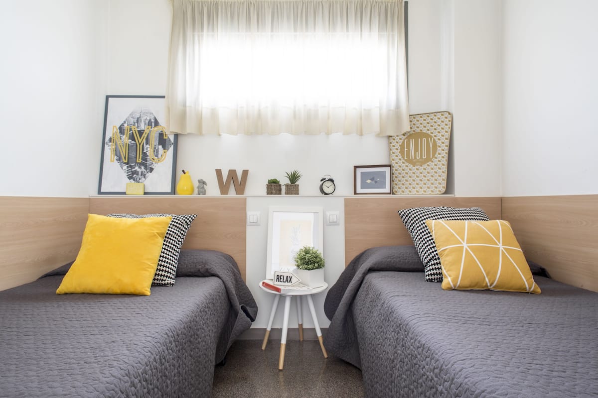 Une chambre d’hôtel avec deux lits, une petite table ornée d’une plante, un tableau encadré et une fenêtre avec des rideaux légers.