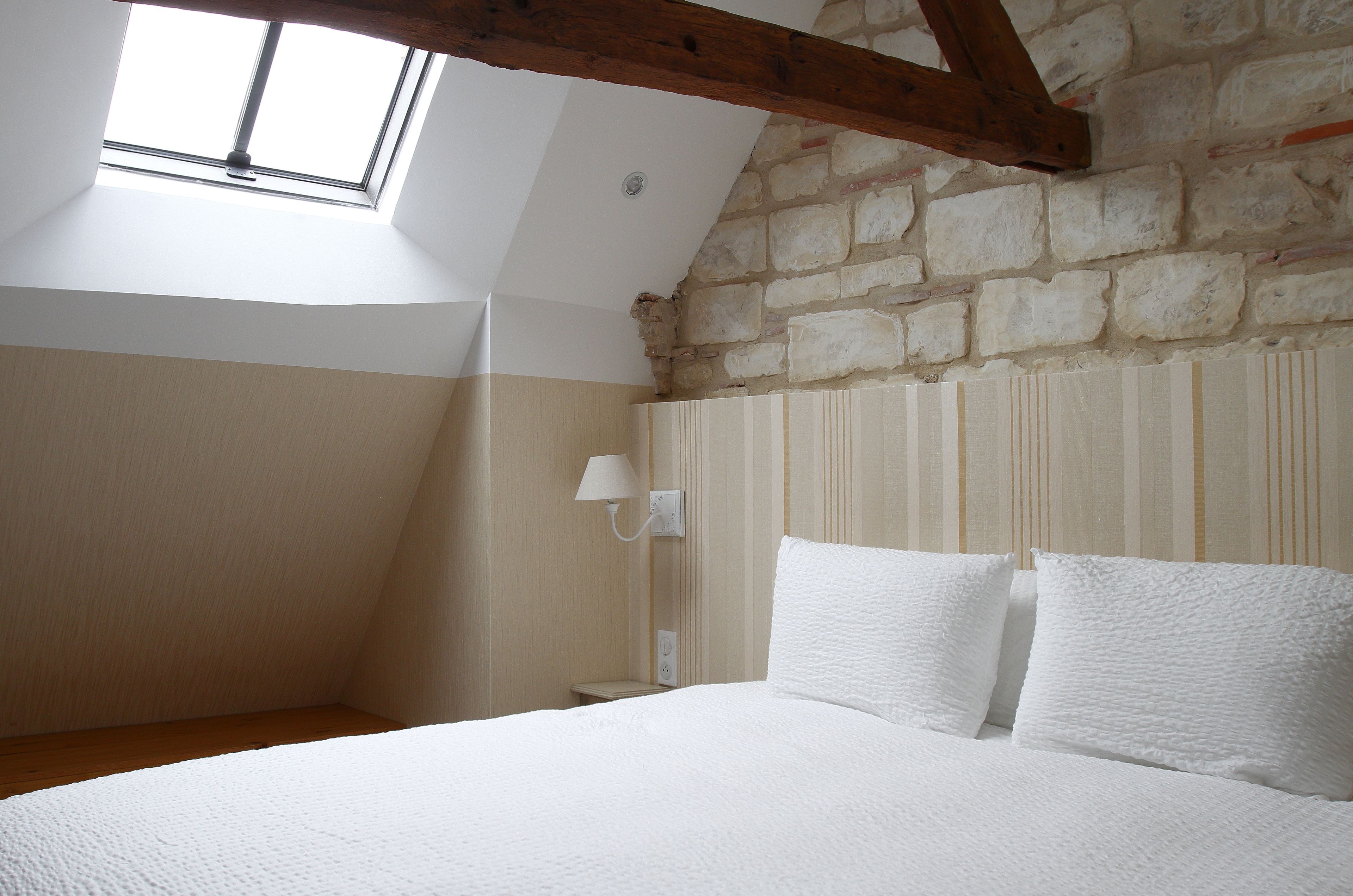 Ein Schlafzimmer mit schrägen Wänden, einem Oberlicht, einem Bett mit weißer Bettwäsche, einem Nachttisch und einer Wandlampe.