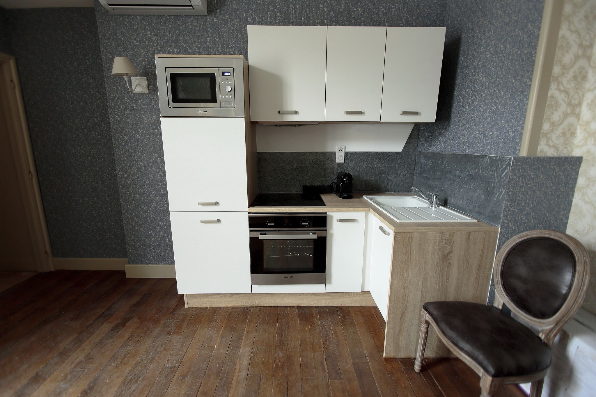 A compact kitchen with a microwave, oven, and sink, wooden flooring, and a chair.