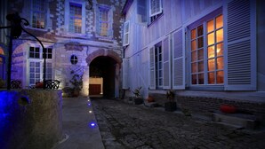 Un patio empedrado con un banco de piedra, una puerta de madera y un arco de ladrillo que da a un edificio con contraventanas blancas y una ventana con luz cálida.