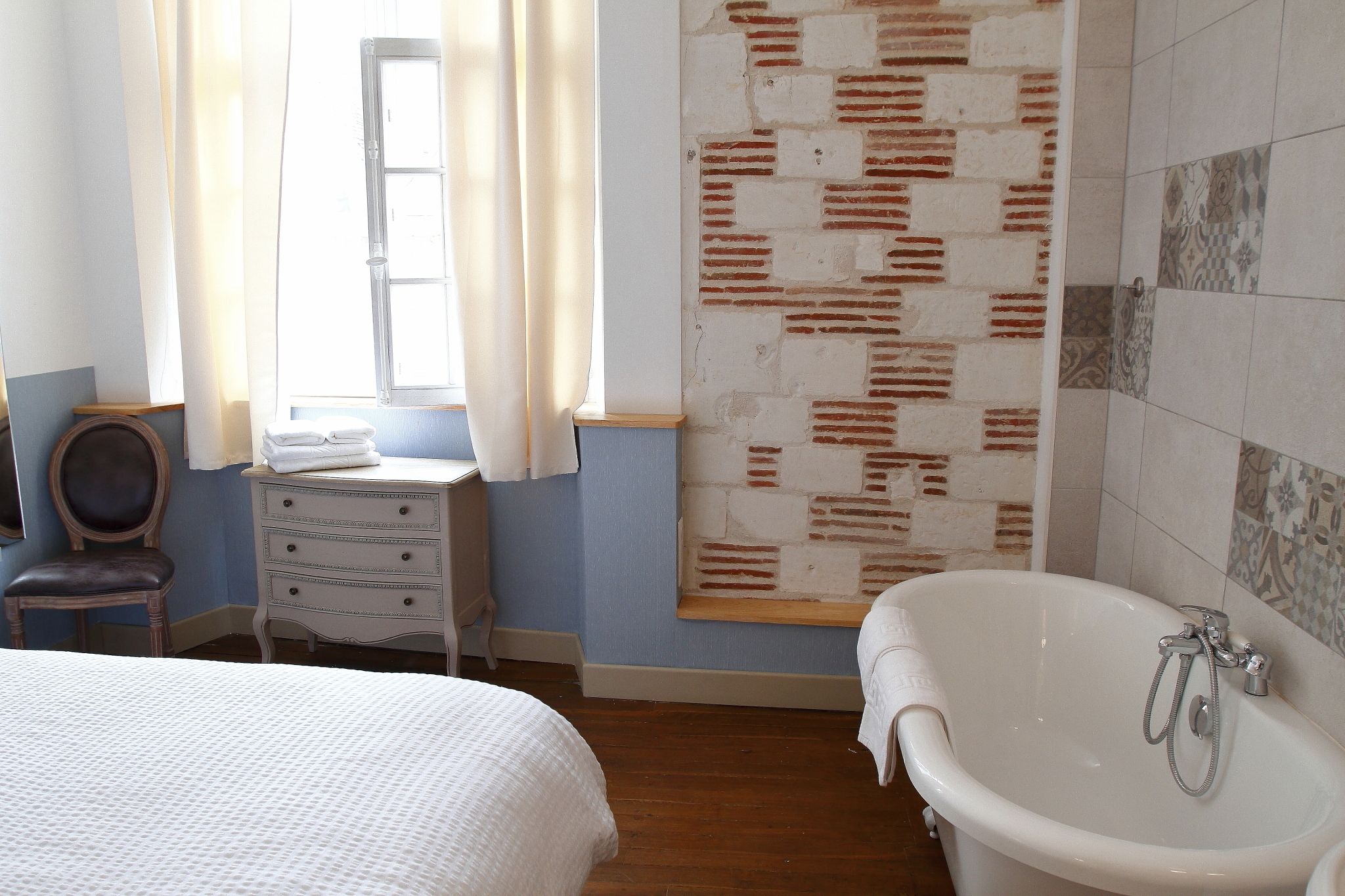 A bedroom with a bed, a dresser, a window with curtains, a brick-patterned wall, and a bathtub.