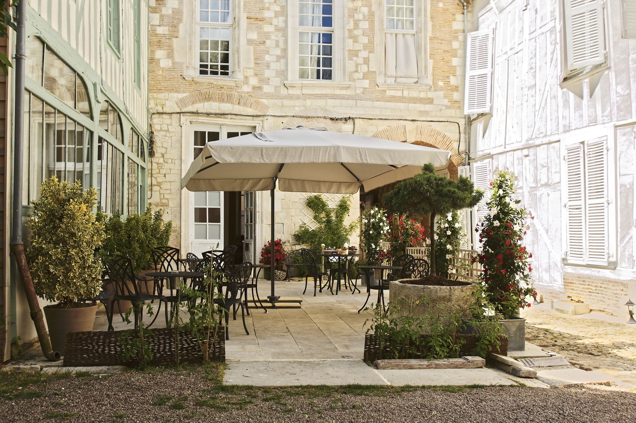 An outdoor patio area with tables and chairs under a large umbrella, surrounded by potted plants and a stone building with shutters.