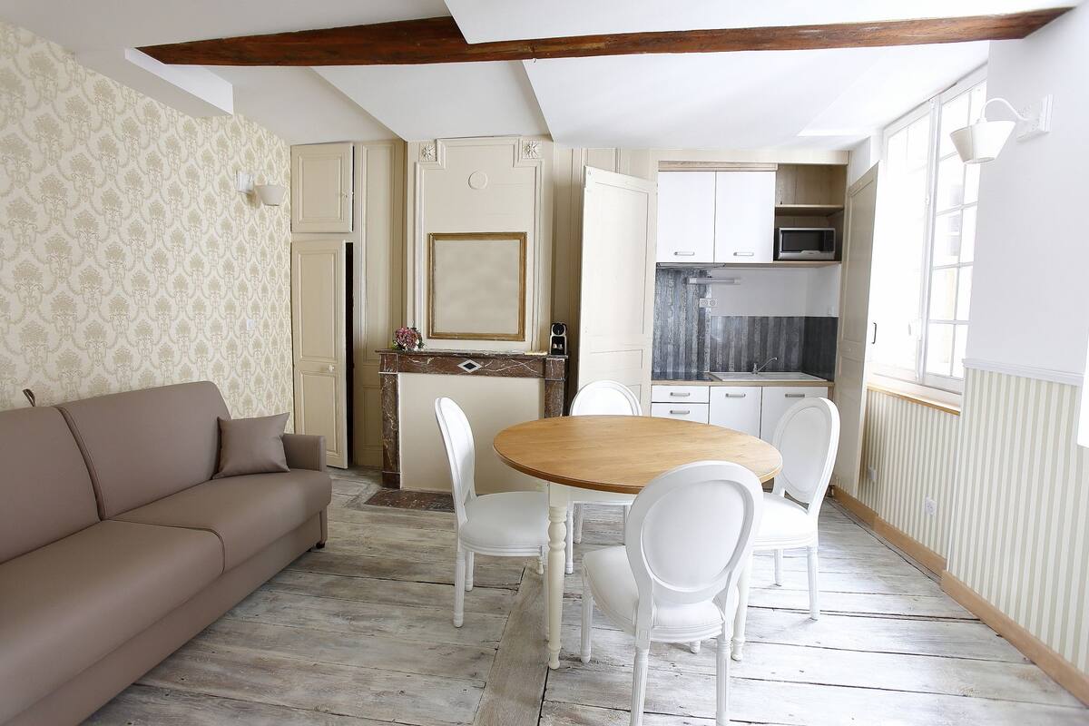 Een gezellige woonkamer met een beige bank, een houten eettafel met witte stoelen, en een keuken met witte kasten en een magnetron.