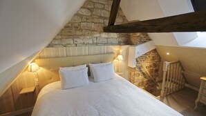 Un dormitorio con techo inclinado, pared de piedra, vigas de madera, una cama con ropa de cama blanca y dos lámparas de noche.