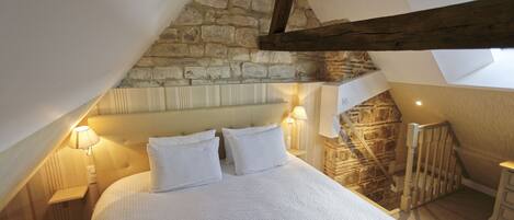Een slaapkamer met een schuine kap, stenen muur, houten balken, een bed met wit beddengoed en twee bedlampjes.