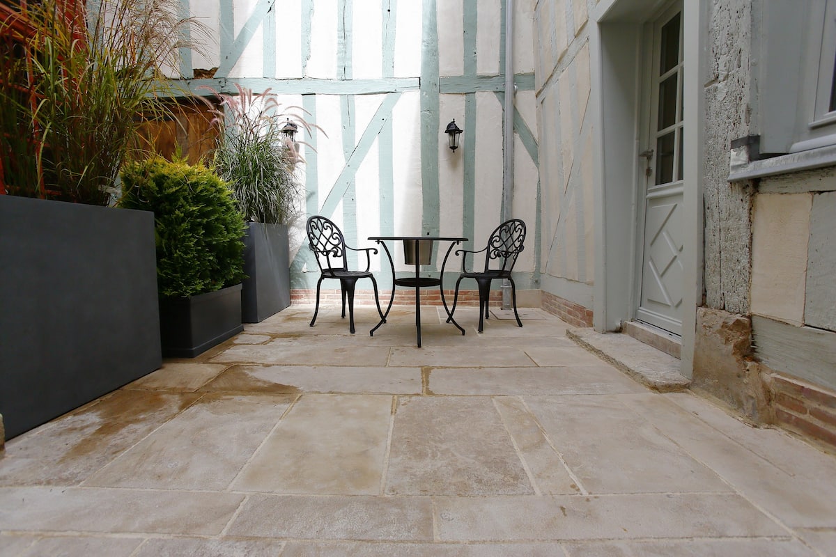 Een binnenplaats met stenen vloer, twee zwarte metalen stoelen, een kleine tafel en plantenpotten.