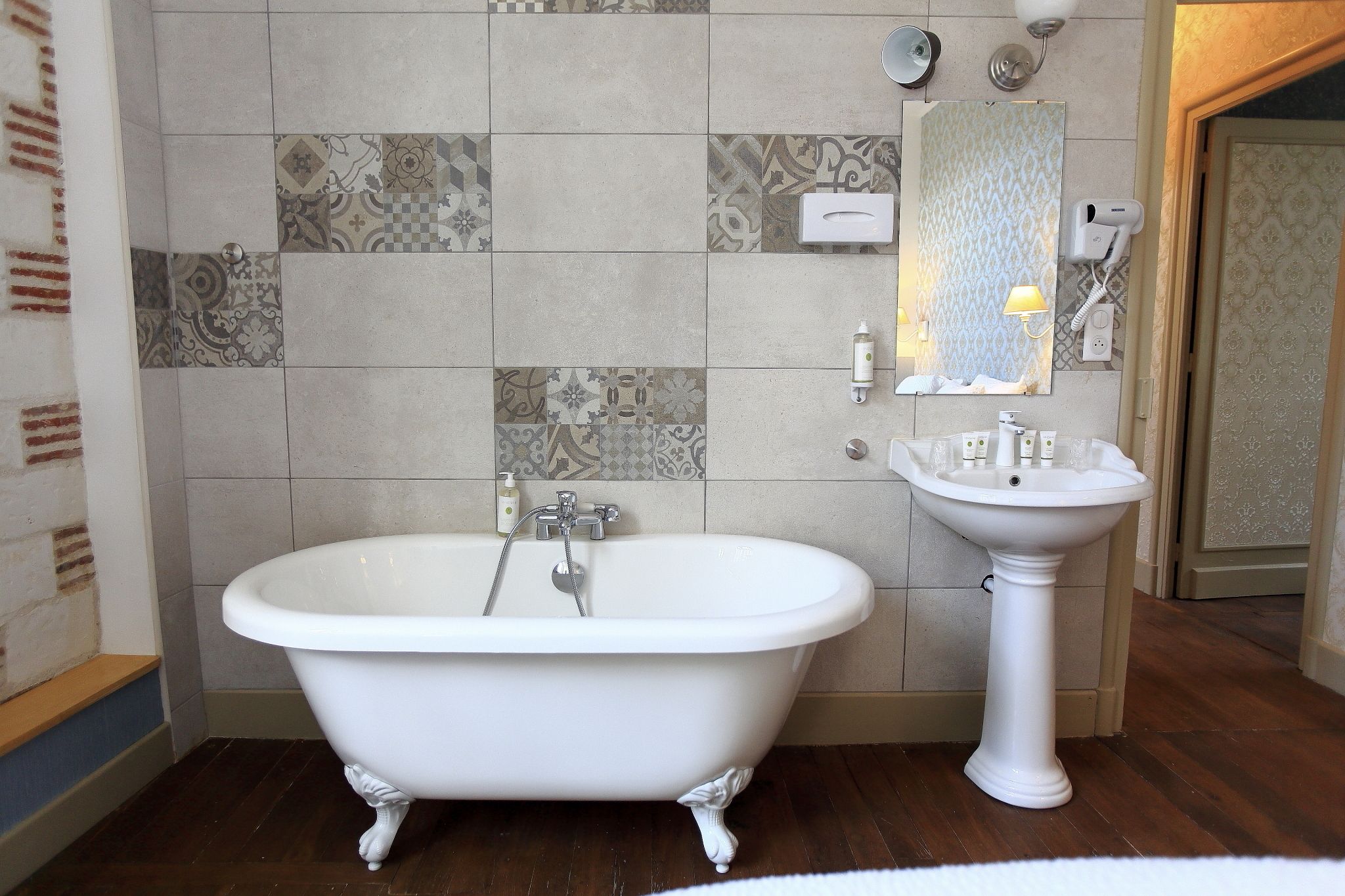A bathroom with a freestanding bathtub, a pedestal sink, and a patterned tile wall.