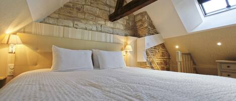 Un dormitorio con pared de piedra, techo inclinado y una cama grande con ropa blanca.