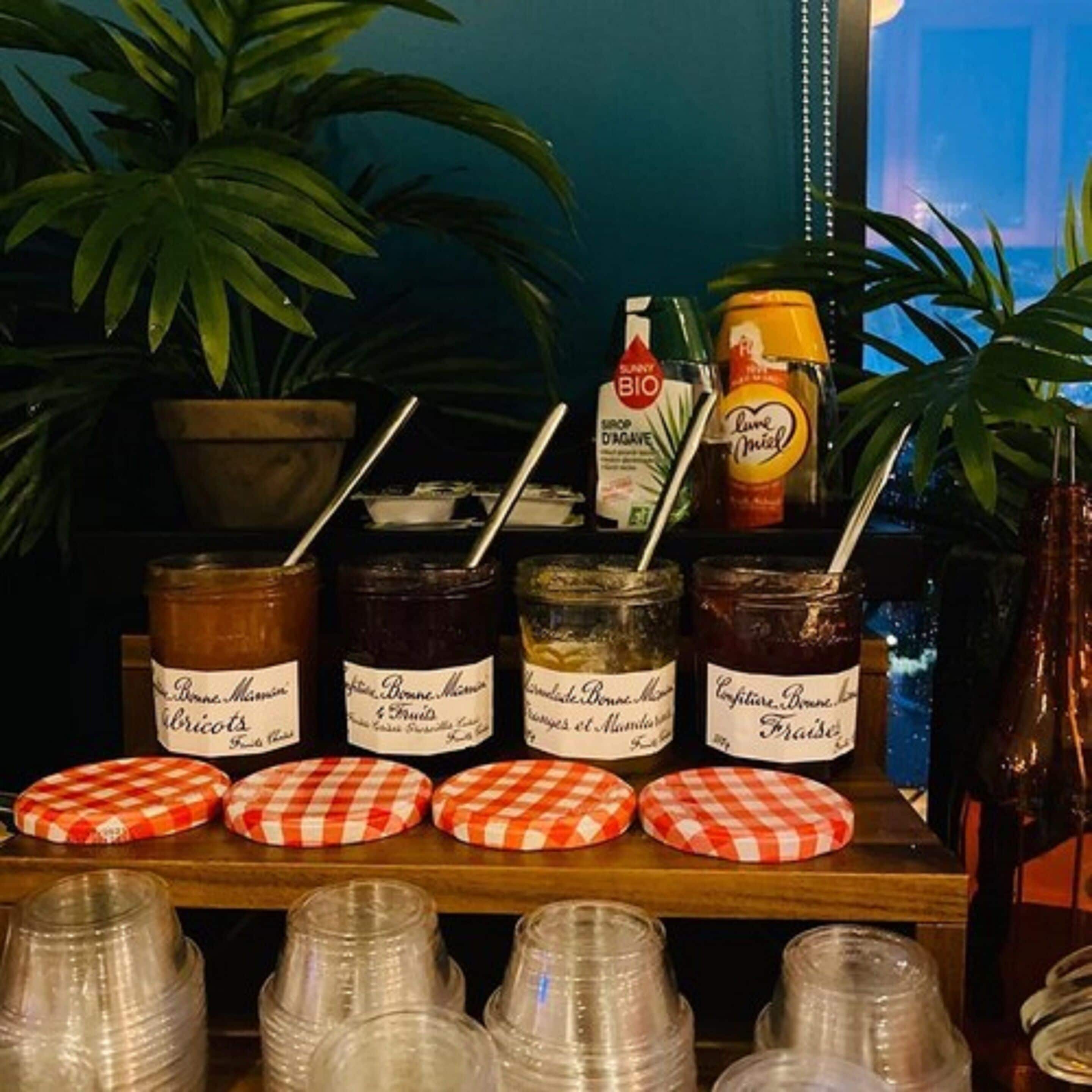 A display of food items and condiments, including jars of preserves and a bottle of juice, with checkered lids and a potted plant in the background.
