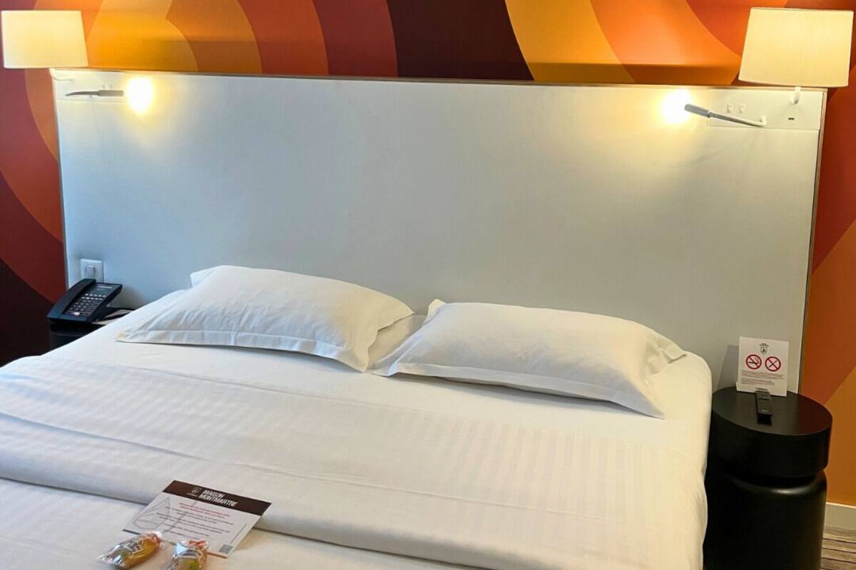 Une chambre d’hôtel avec un lit, du linge de lit blanc, deux oreillers, une table de chevet avec un téléphone, un panneau indiquant une interdiction, et un mur orné d’un motif abstrait et coloré.
