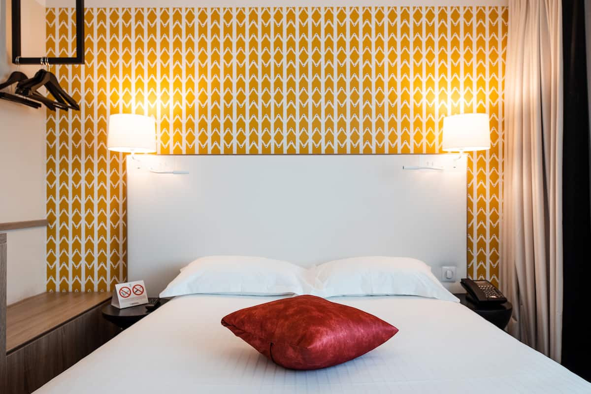 Une chambre d’hôtel avec un lit, deux lampes de chevet, un oreiller rouge, une table de chevet avec un téléphone et un mur recouvert d’un papier peint à motifs.
