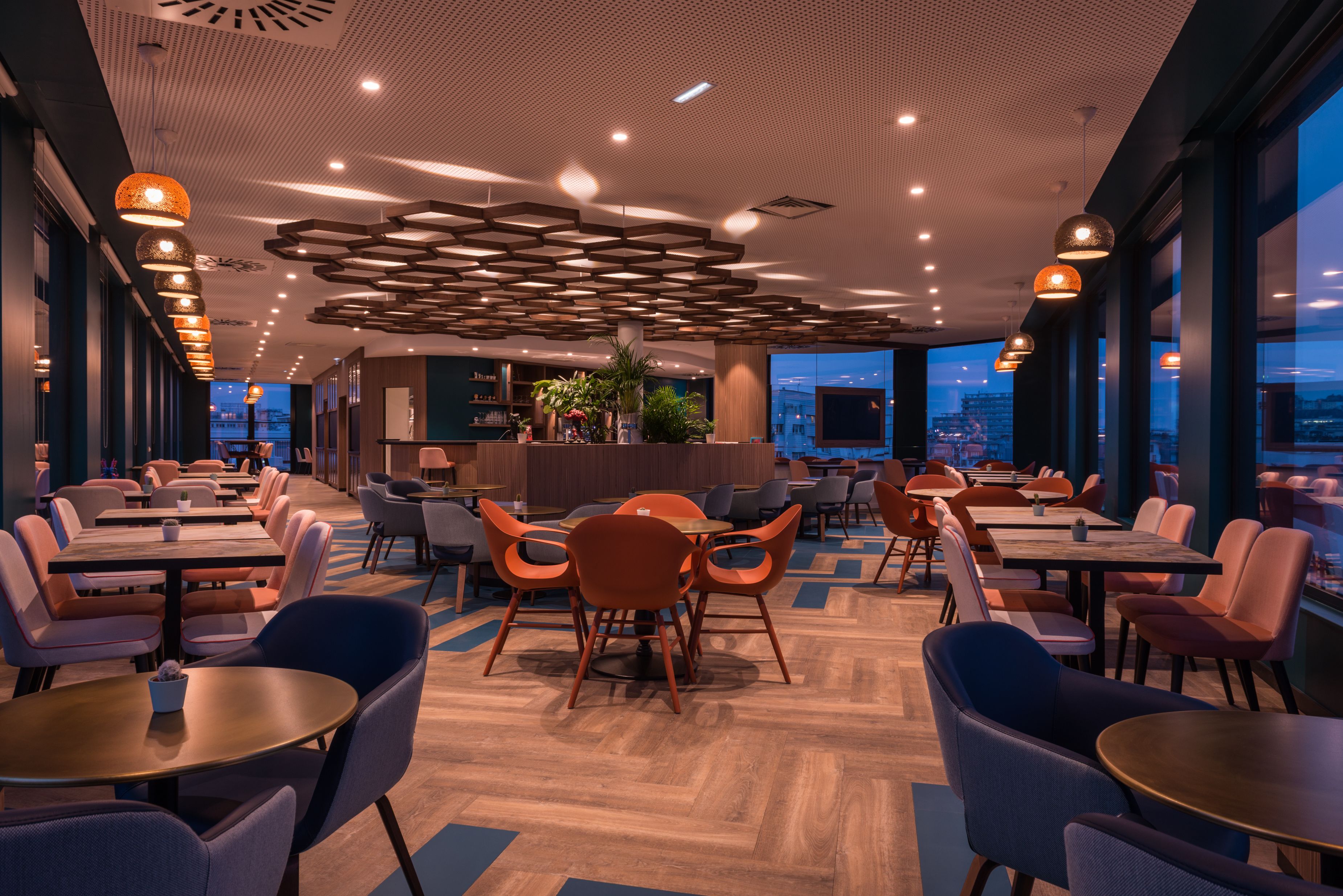 Un restaurant moderne avec un plafond haut, un plancher en bois et des chaises orange. Les tables sont disposées au centre, avec un bar et des plantes en arrière-plan.