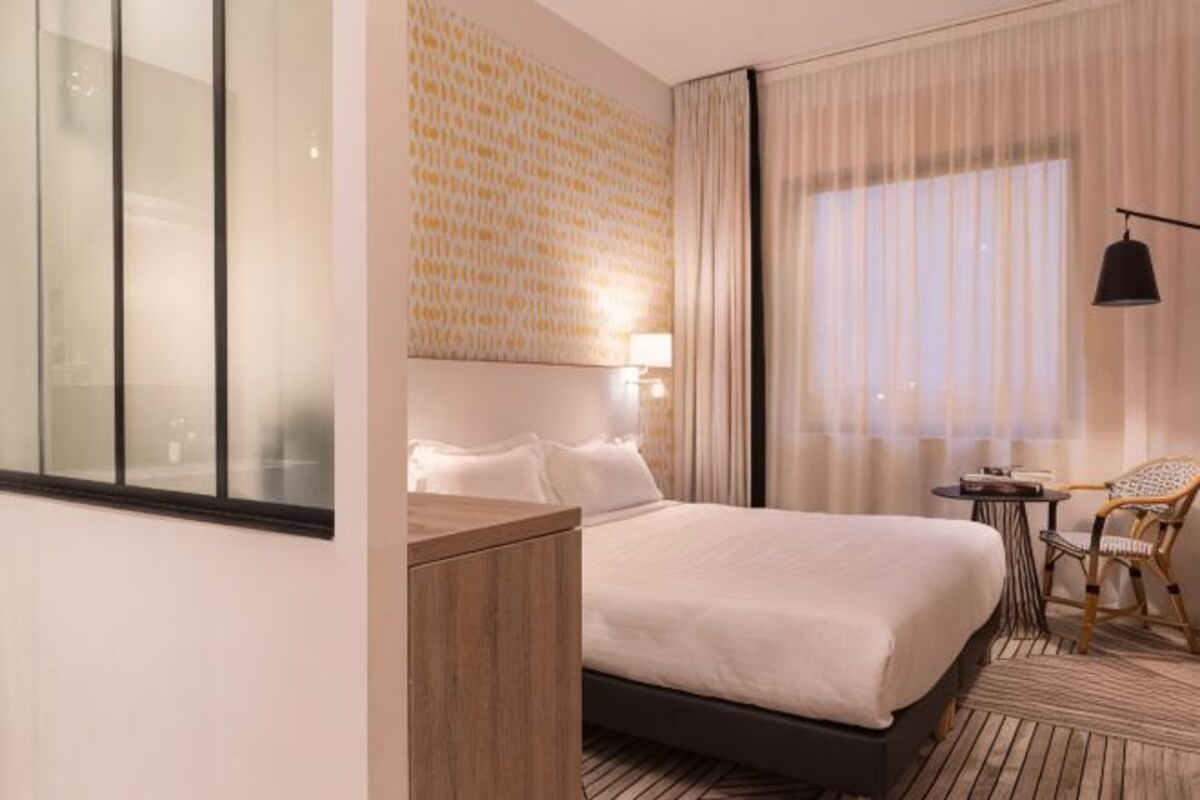 Hotelkamer met een groot bed, een houten nachtkastje, een kleine tafel met een stoel en een groot raam met gordijnen.