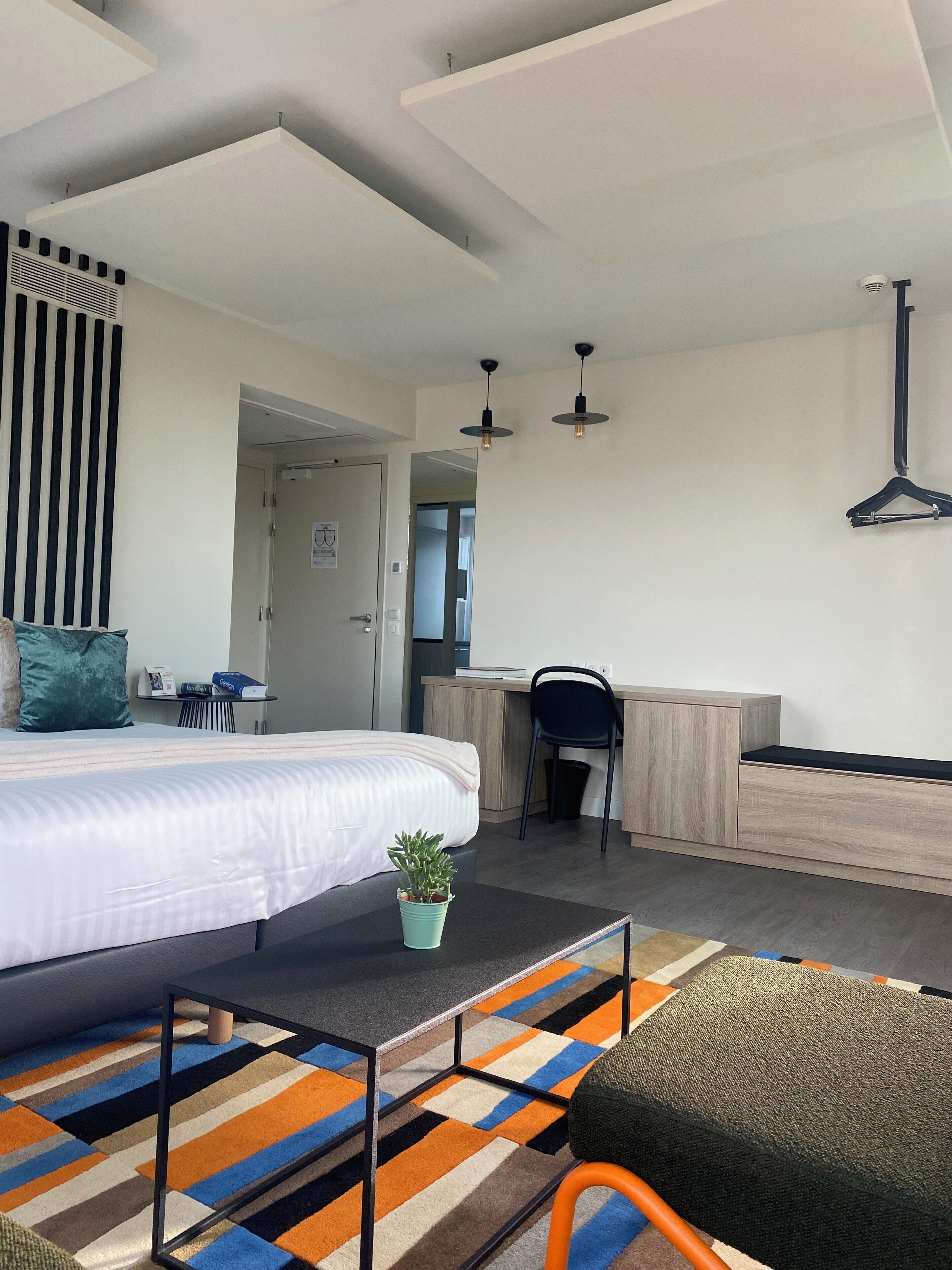 A modern hotel room with a bed, a desk, a chair, a small table, a hanging coat rack, and a colorful striped rug.