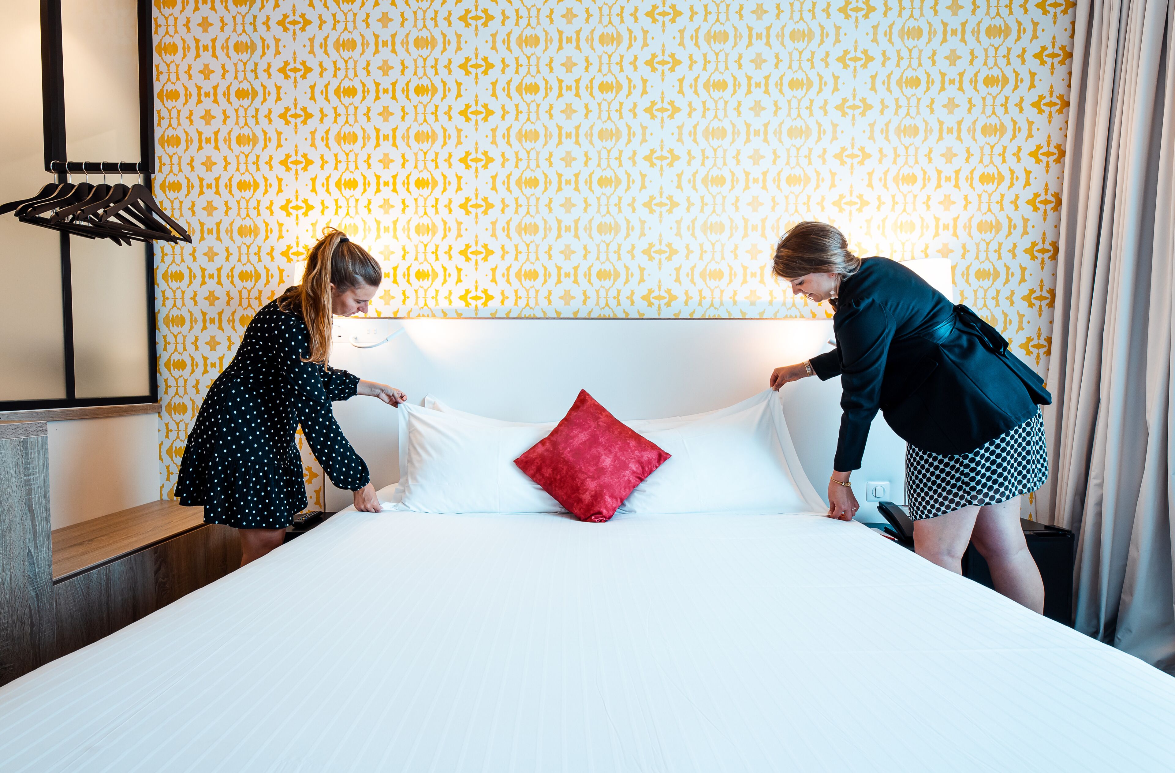 A hotel room with a patterned wallpaper, a neatly made bed with white and red pillows, and a wooden nightstand.