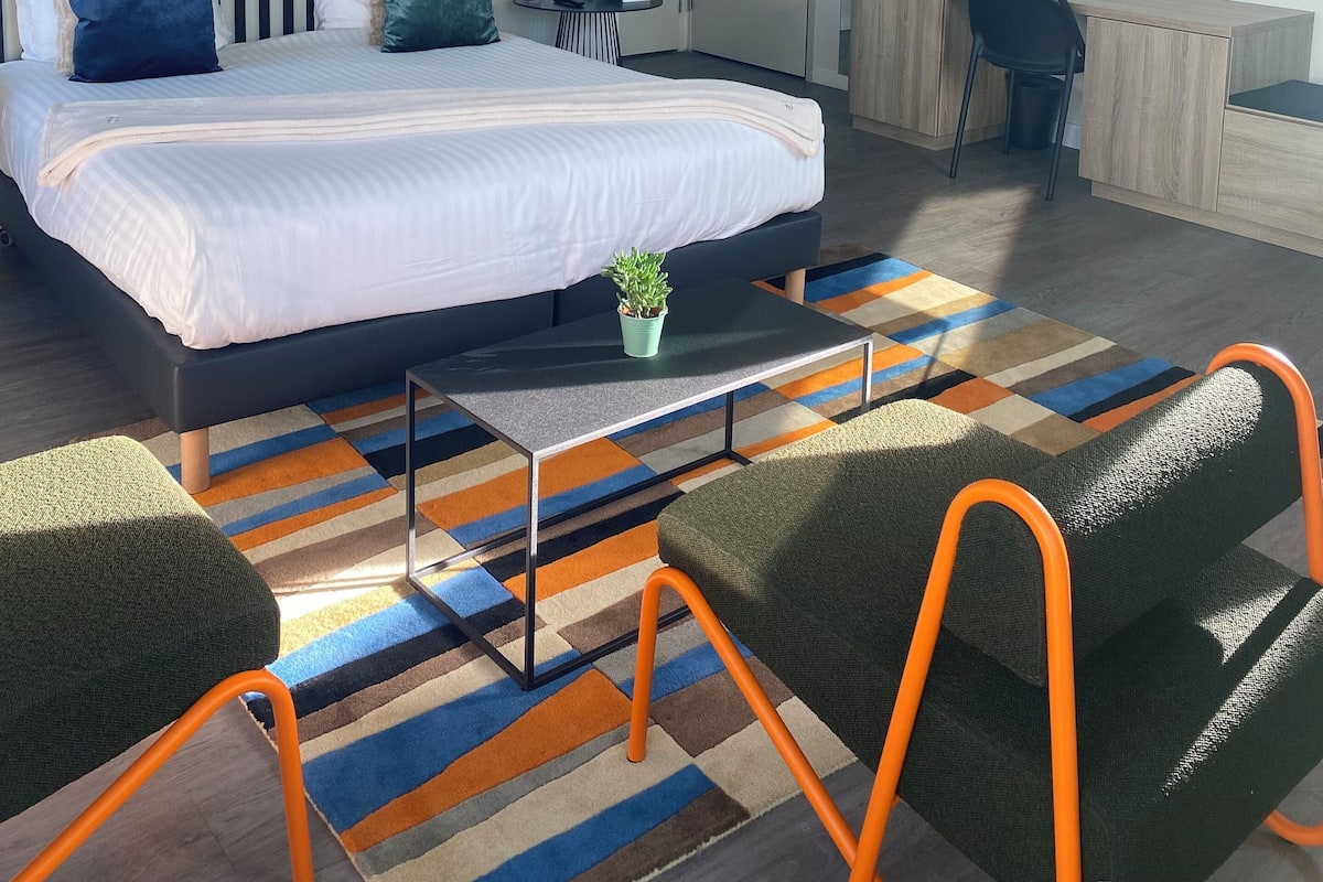Une chambre d’hôtel moderne avec un grand lit, un bureau et une chaise. On y trouve une petite table avec une plante, ainsi qu’un tapis rayé et coloré sur le sol.