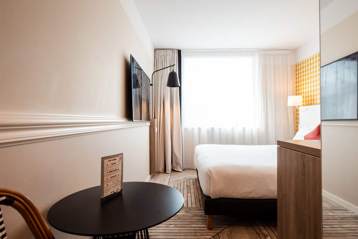 Une chambre d’hôtel avec un lit, une petite table ronde, une chaise, une lampe et un téléviseur fixé au mur.