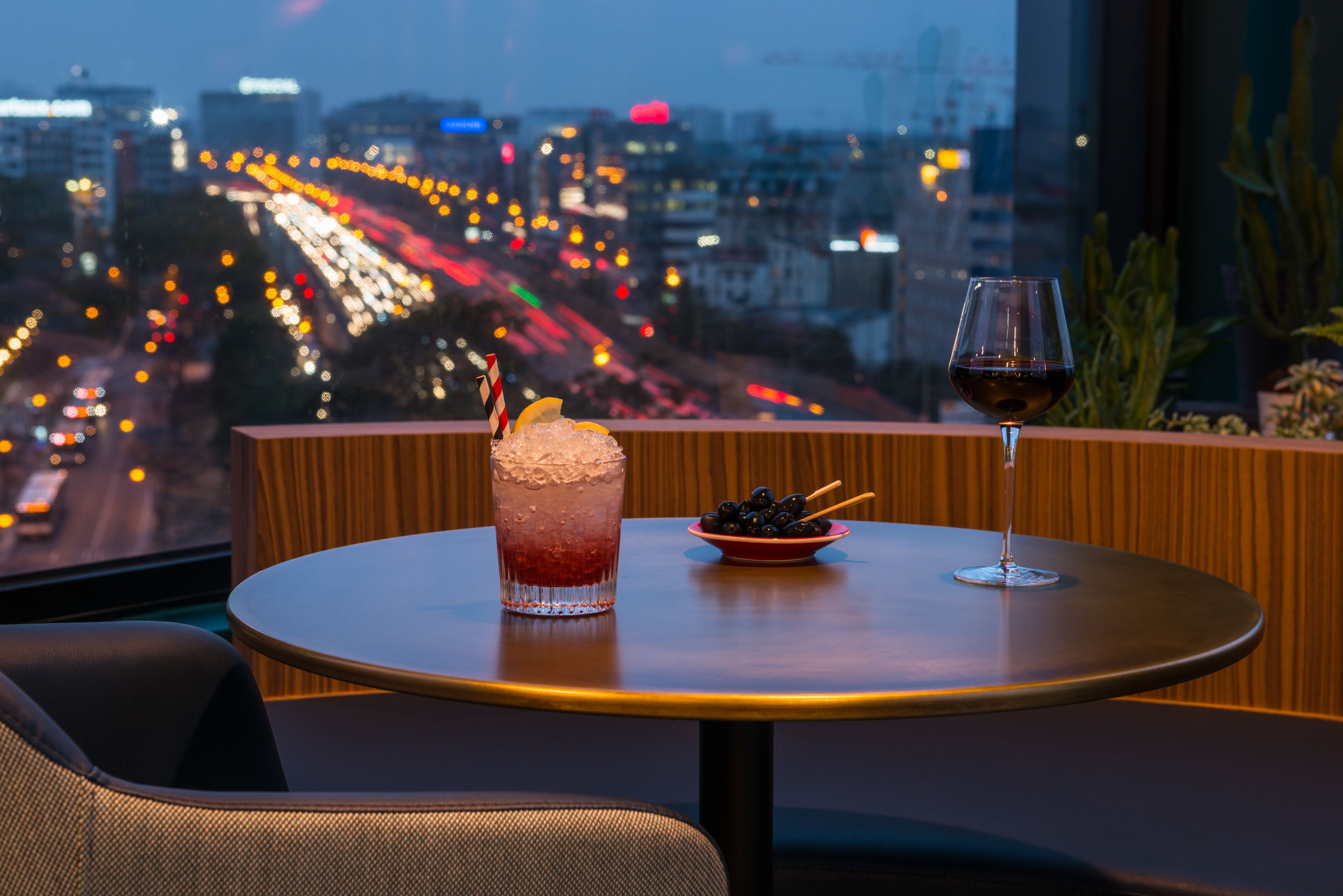 Een tafel met een drankje en een schaal olijven, een stoel, en een stadszicht door een raam.