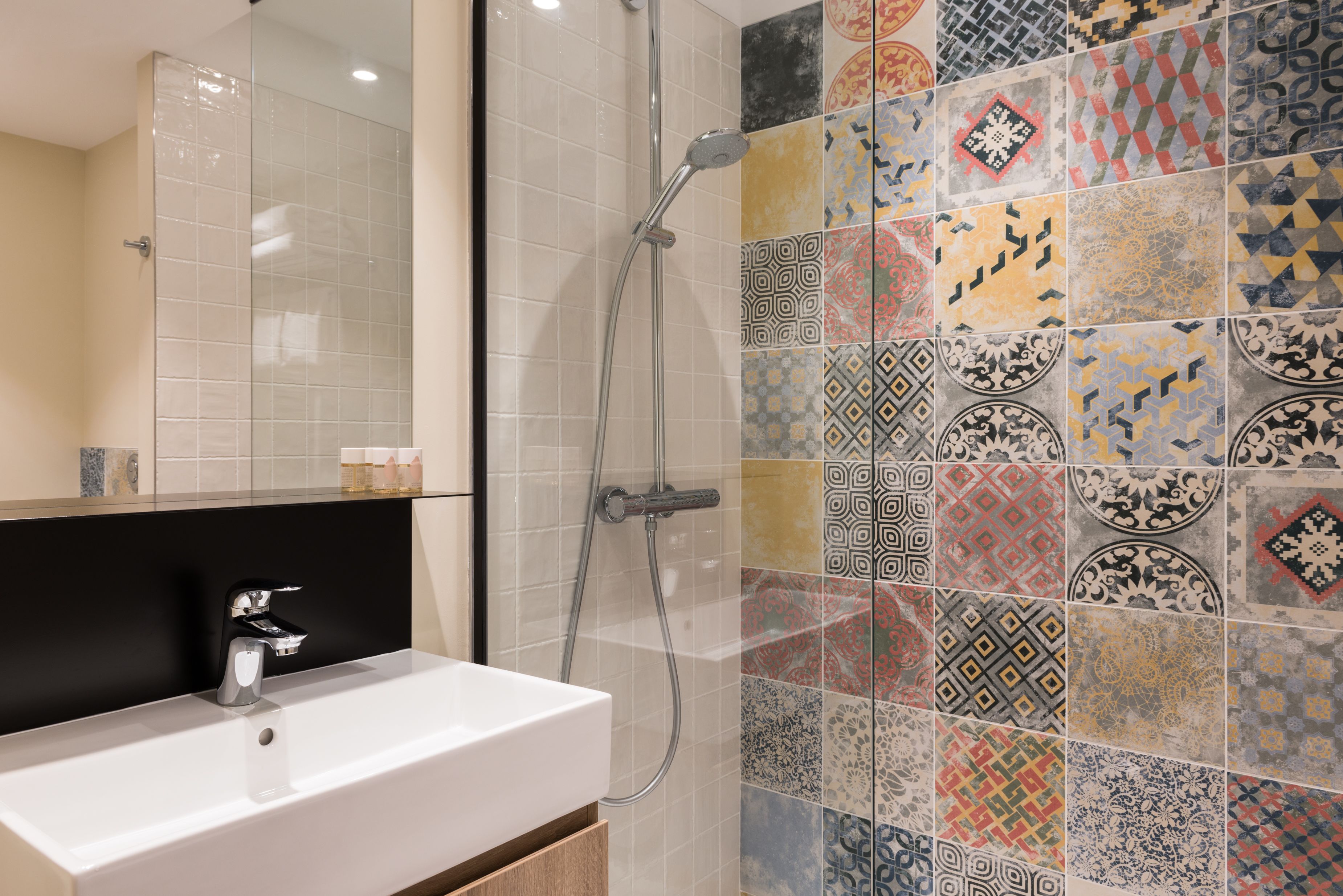 Un baño moderno con lavamanos blanco, cabina de ducha con vidrio y pared de azulejos con distintos diseños.