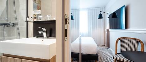 Ein modernes Hotelzimmer mit einem großen Badezimmerwaschbecken, einer Dusche, einem Bett mit weißer Bettwäsche, Holzboden, einem Esstisch mit Stuhl und einem an der Wand befestigten Flachbildfernseher.