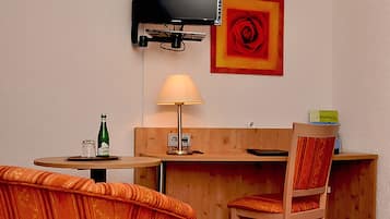 A hotel room with a TV mounted on the wall, a painting of a rose, a lamp, a bottle of water, and a chair.