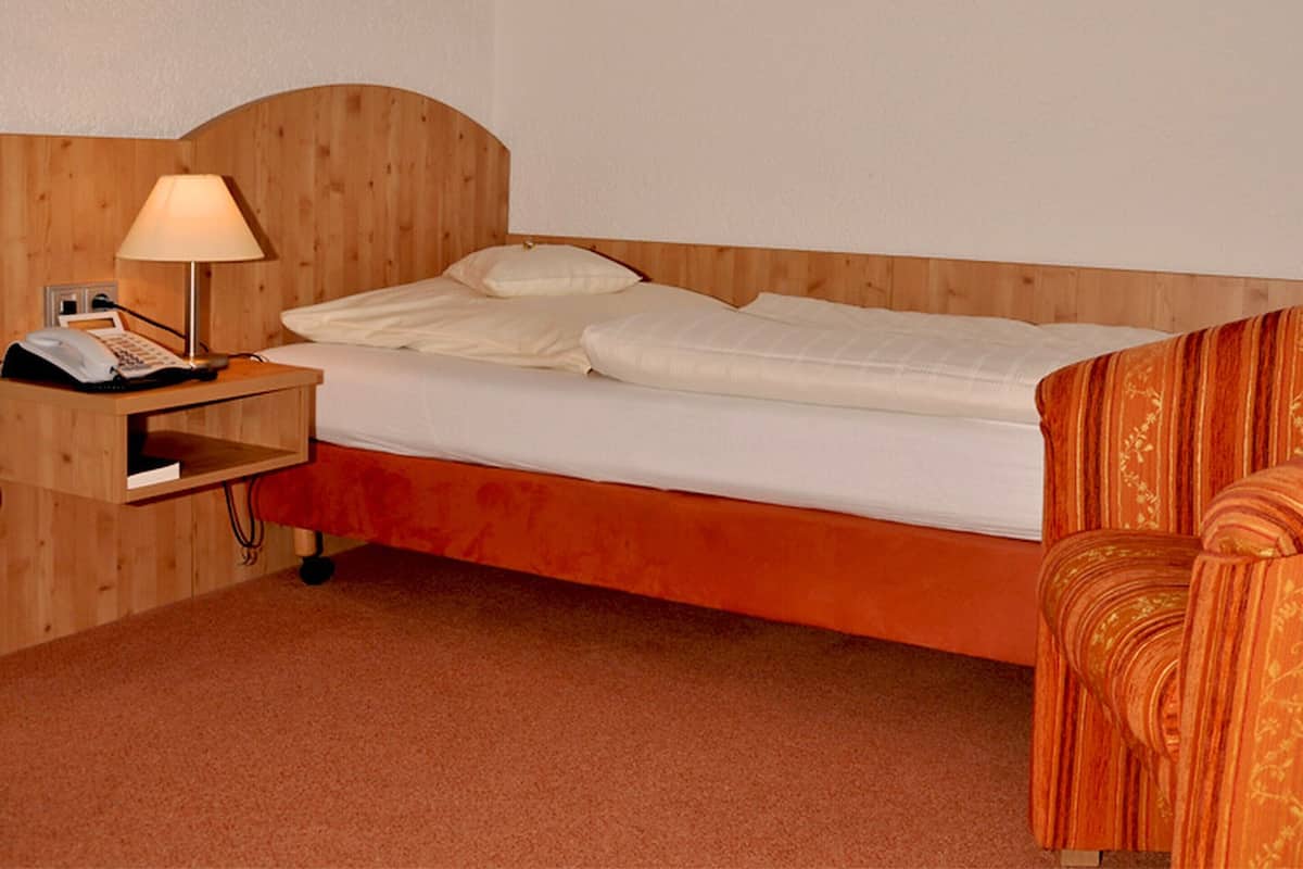 Habitación de hotel con cabecera de madera, cama individual con ropa de cama blanca, mesita de noche con lámpara, teléfono y silla con cojín a rayas.