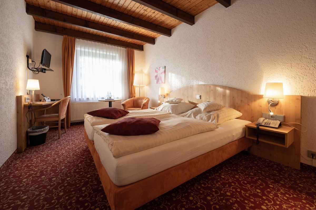 Una habitación de hotel con una cama grande, dos mesitas de noche con lámparas, un escritorio con una silla, una pequeña mesa con una silla y una ventana con cortinas.
