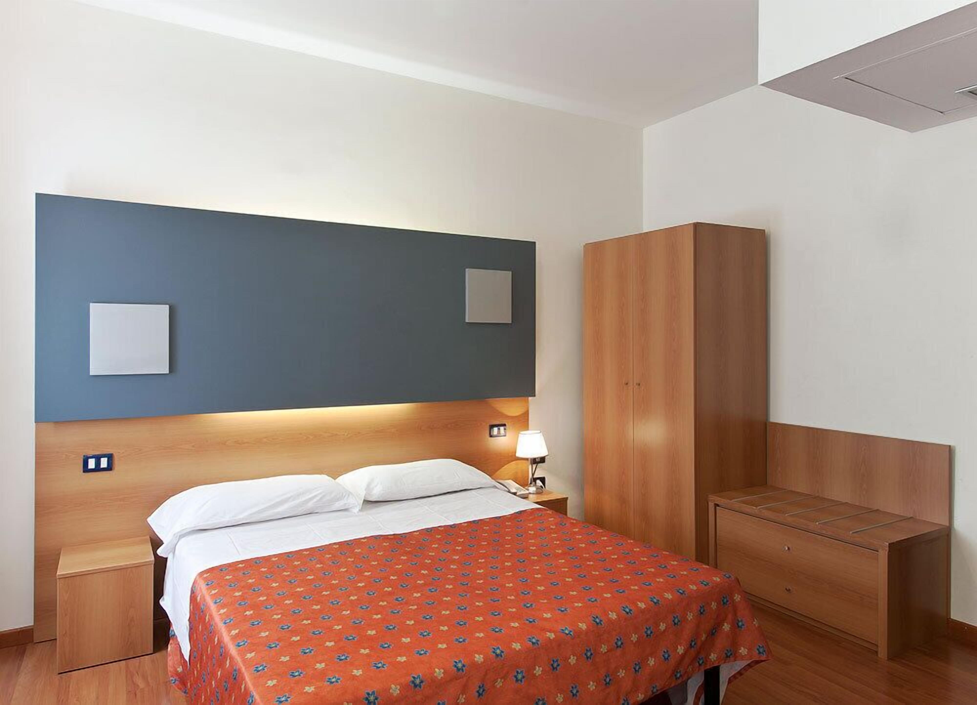 Une chambre d’hôtel comprenant un lit, une table de chevet, une lampe de chevet, une armoire et un petit bureau.
