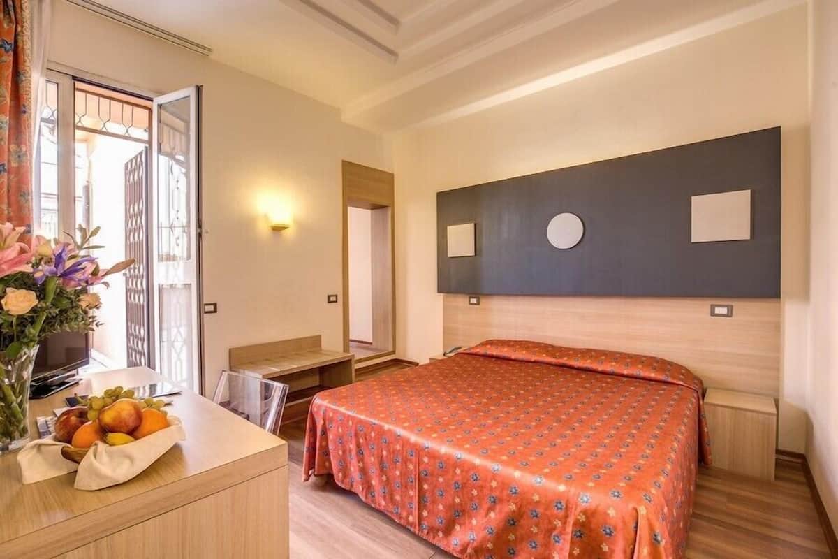 A hotel room with a bed, a desk with a vase of flowers, and a wall-mounted TV.