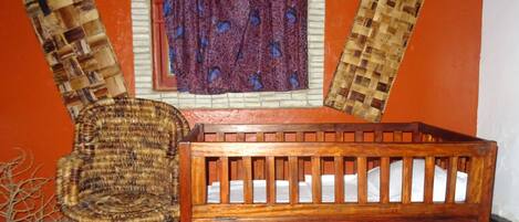 Una cuna de madera con una cesta tejida a su lado, una ventana con cortina morada y una pared naranja.