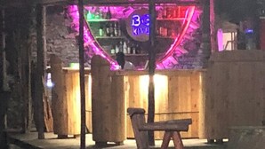 Un área de bar al aire libre con un letrero de neón, mobiliario de madera y una pared de piedra.