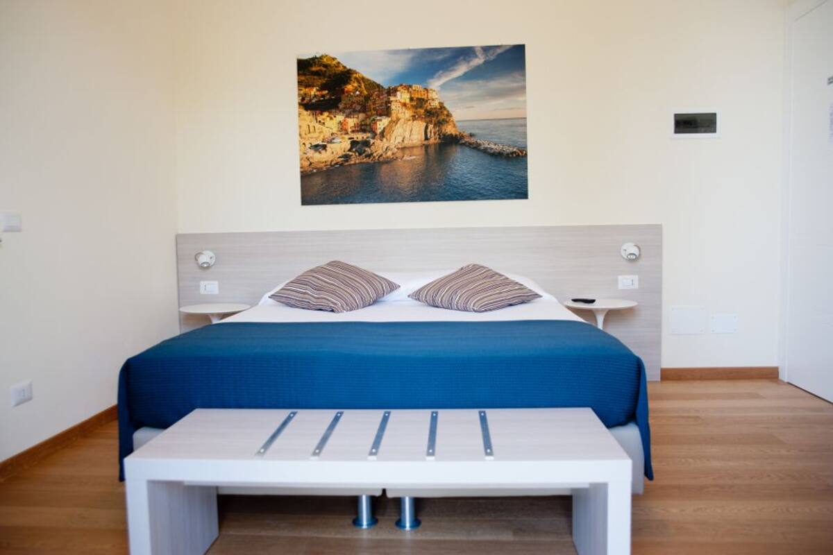 Un dormitorio con cama, almohadas, mesitas de noche, un banco y un cuadro de una escena costera.