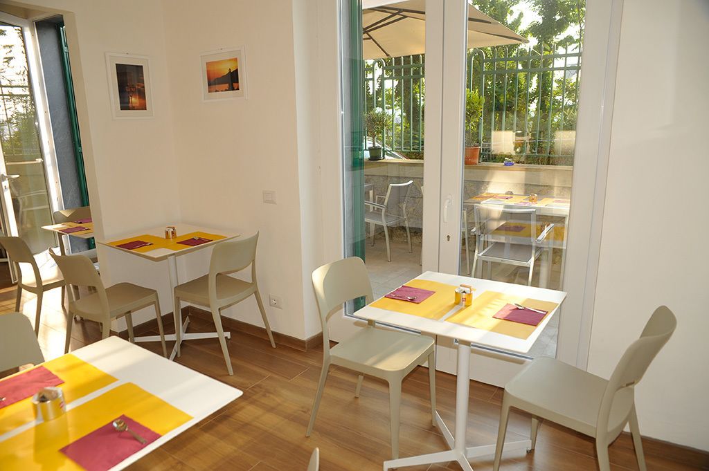 Un comedor luminoso con mesas y sillas blancas, individuales amarillas y vistas a un patio exterior con sombrillas.