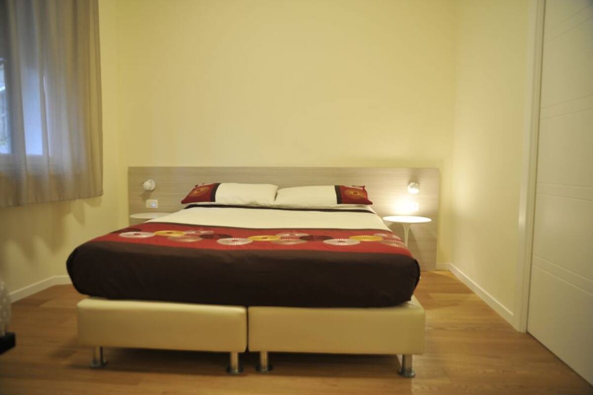 Una cama bien hecha con una colcha estampada, una mesita de noche con una lámpara y una ventana con cortinas.