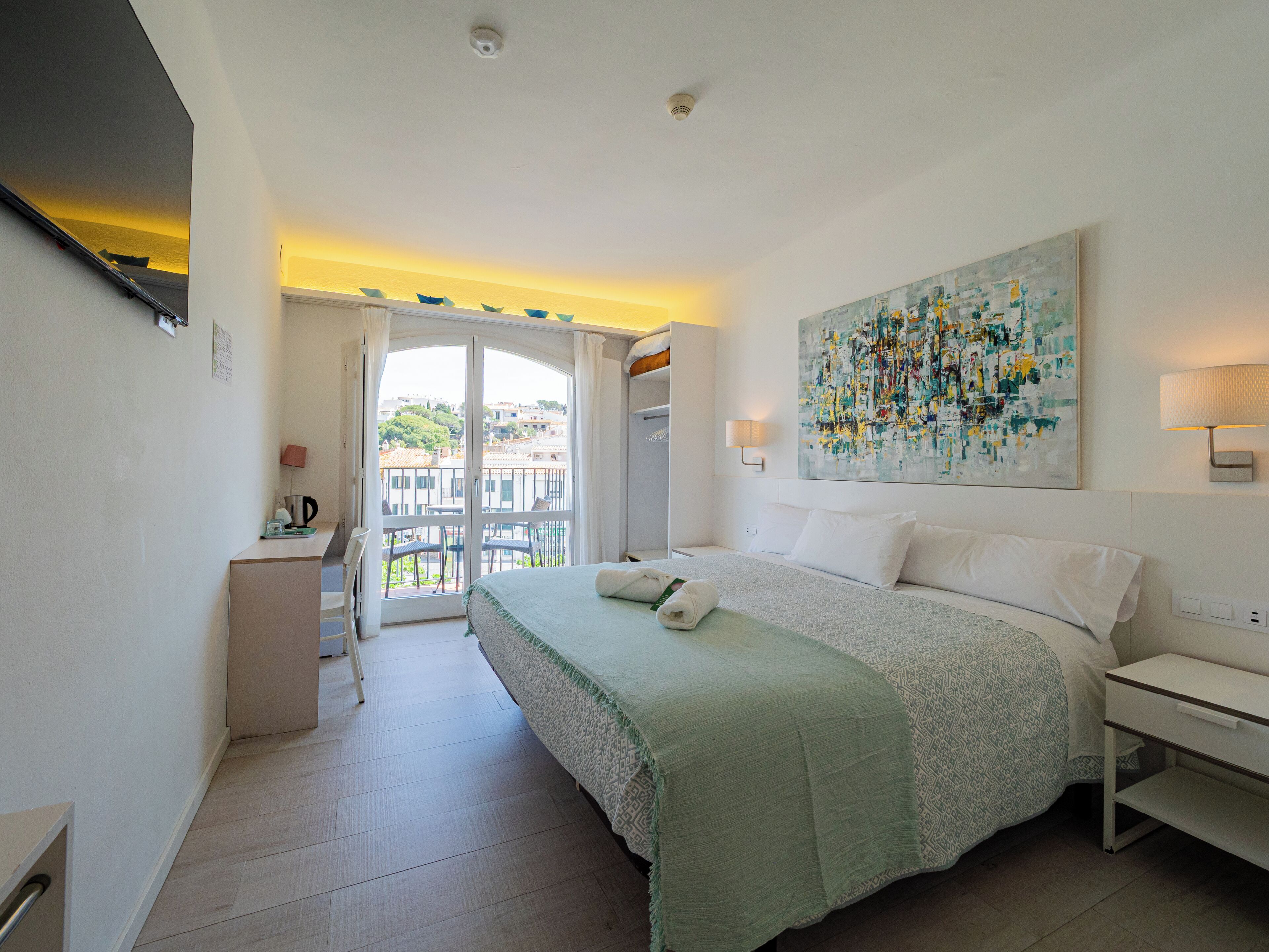 A hotel room with a bed, a desk, a chair, a painting, and a balcony with a view of the city.
