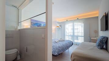 A hotel room with a bathroom, a bed with a patterned bedspread, a bedside table, a chair, and a window with a view of the sea.