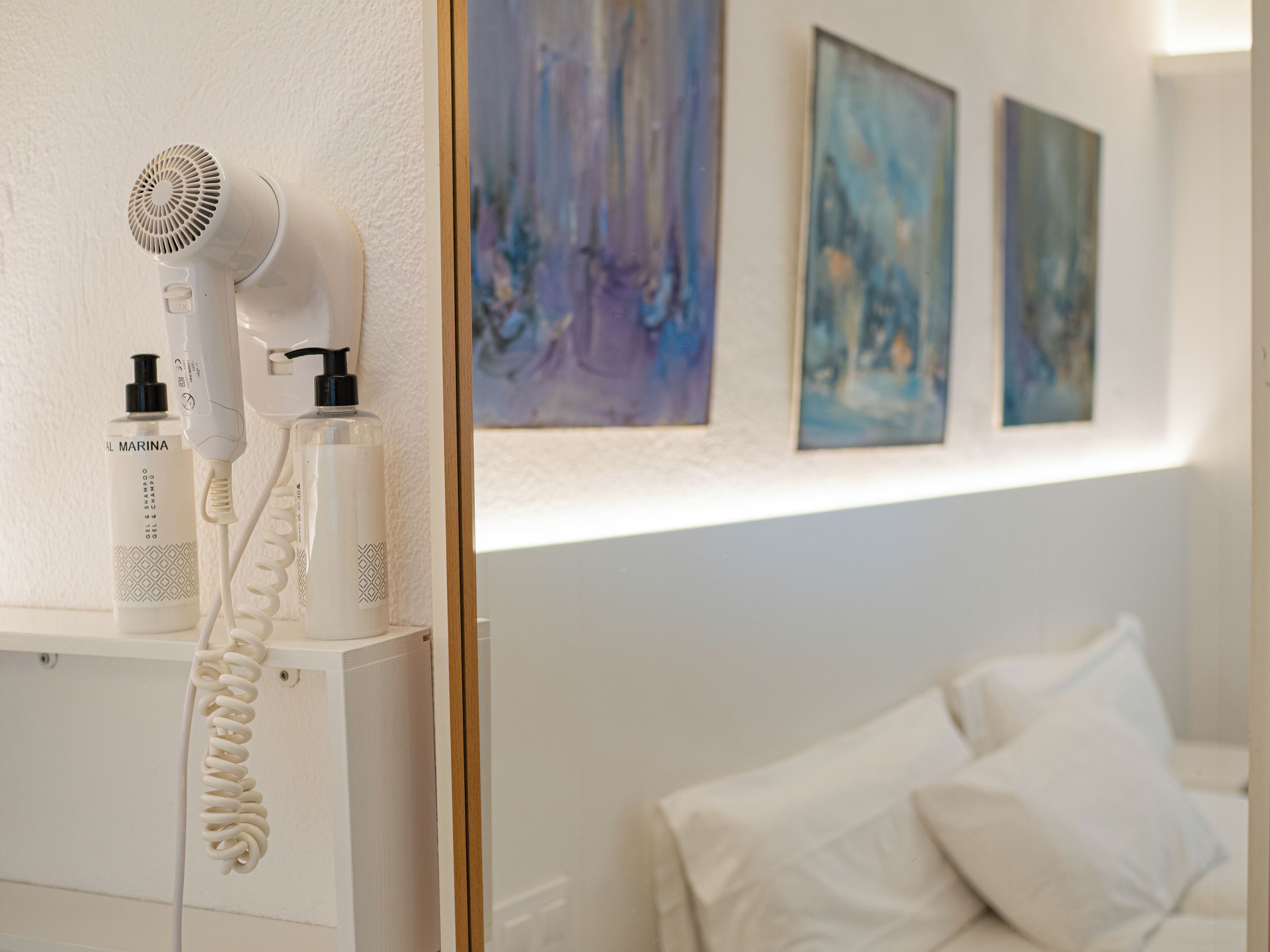 Un secador de pelo y una botella de producto capilar sobre un estante blanco en una habitación con cama y pinturas abstractas en la pared.