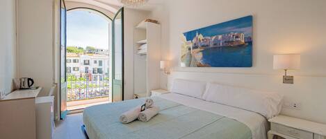 A hotel room with a large bed, a painting of a coastal town, a balcony with a view of buildings, and a unique chandelier.