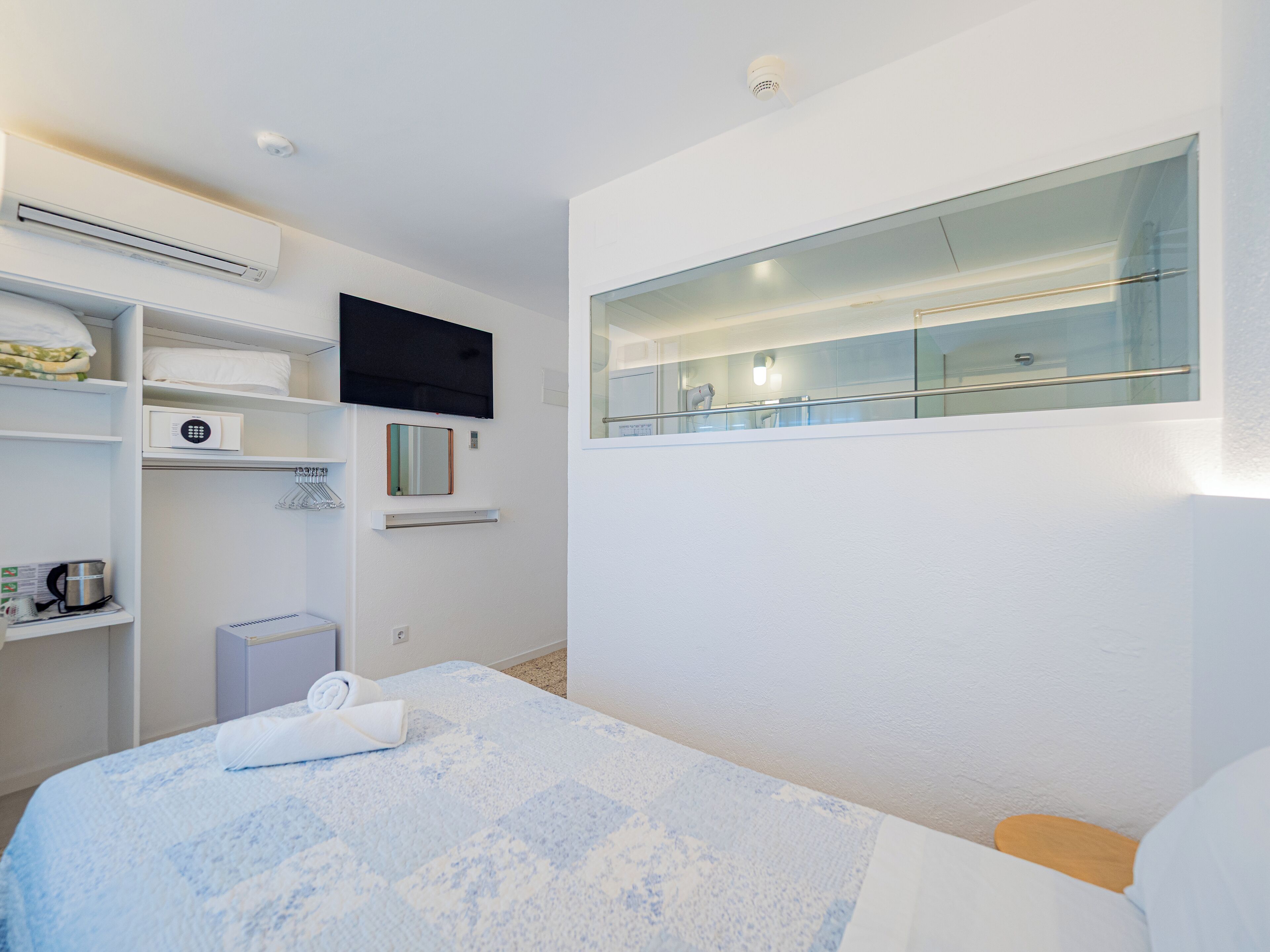 A modern hotel room with a bed, a TV mounted on the wall, a small shelf with toiletries, and a window with a view of the outside.