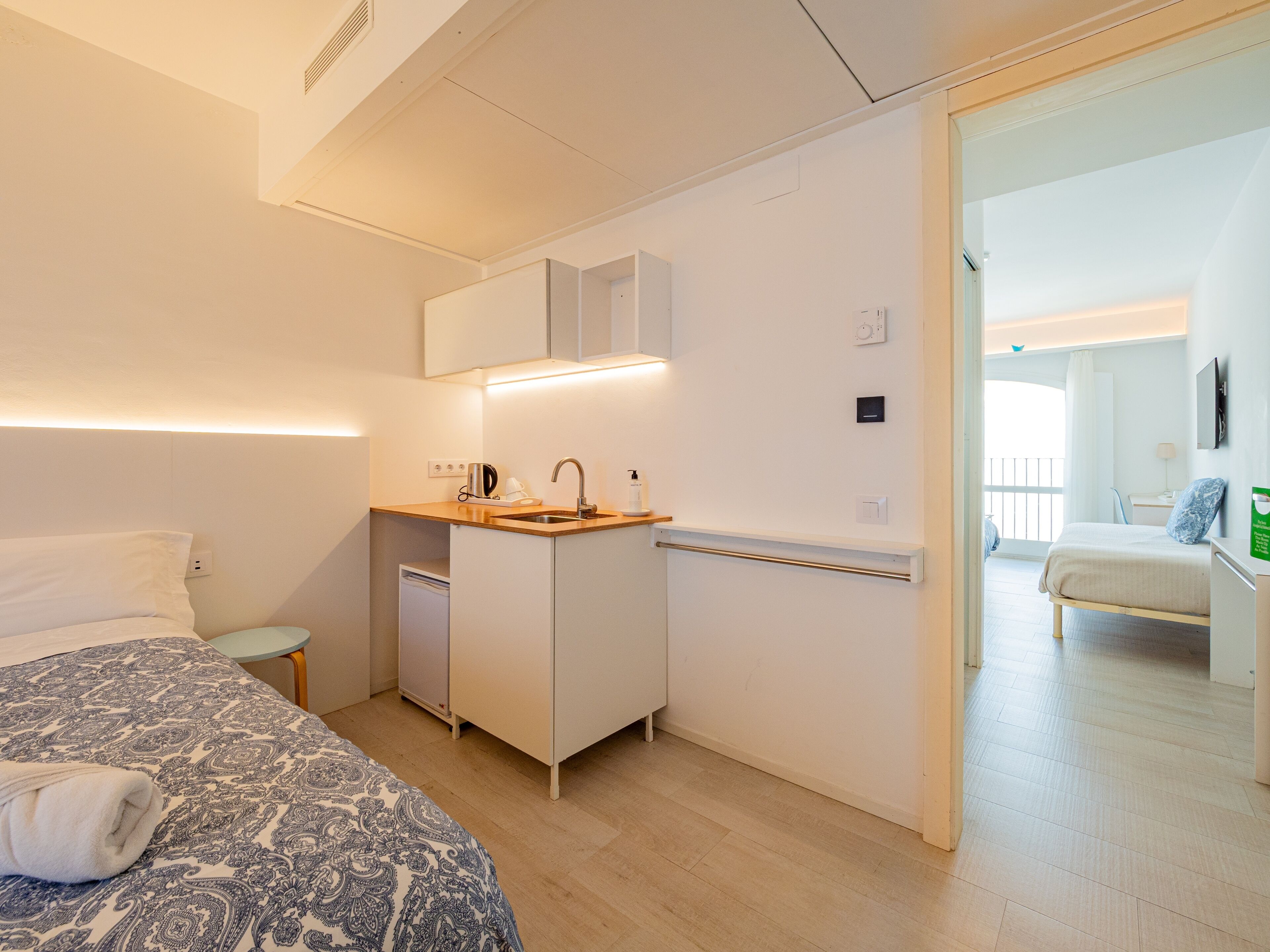 A modern hotel room with a bed, a small kitchenette, a bathroom, and a view of the balcony.