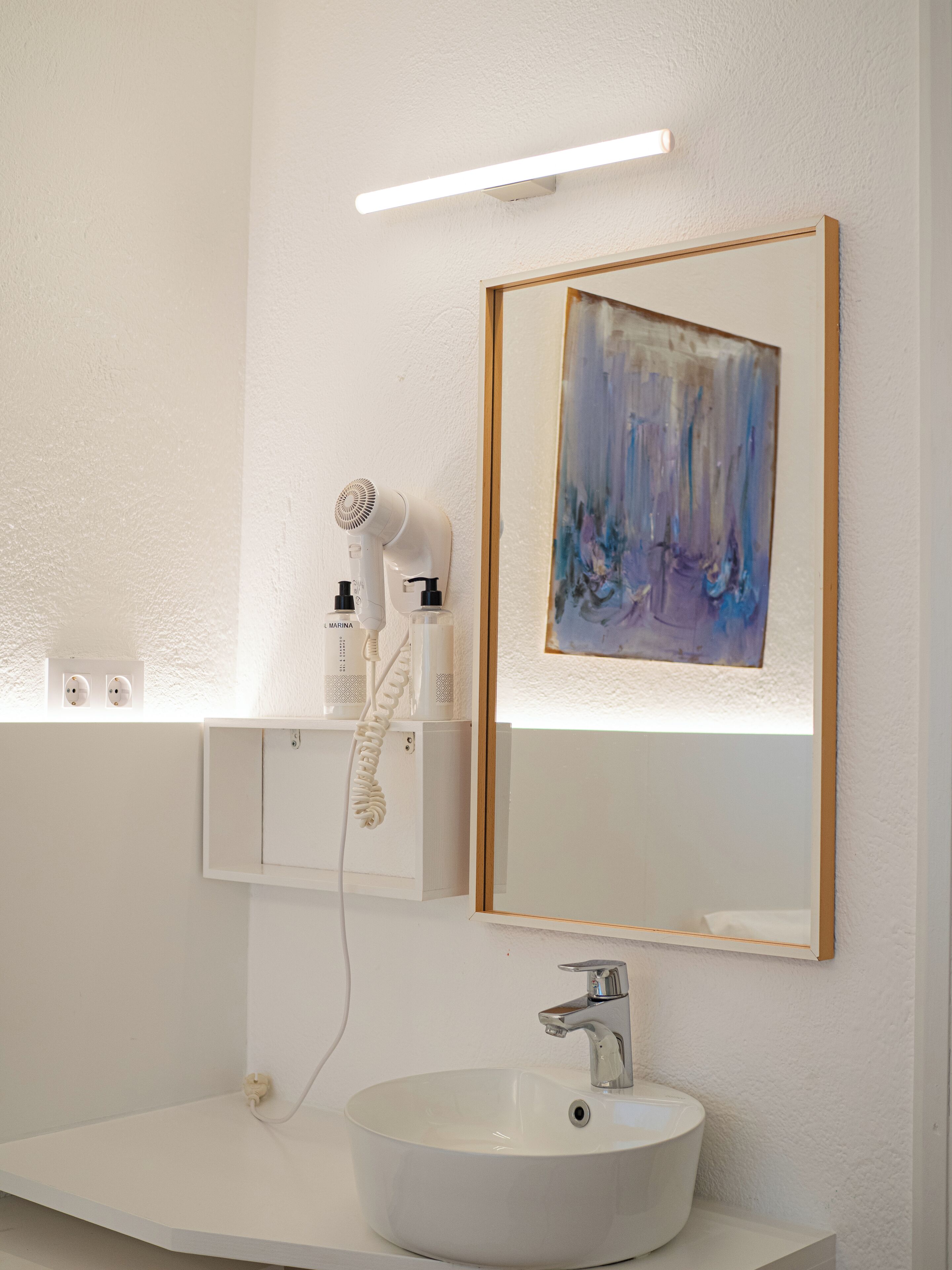 Une salle de bain avec un lavabo blanc, un miroir, un sèche-cheveux et un tableau abstrait encadré.