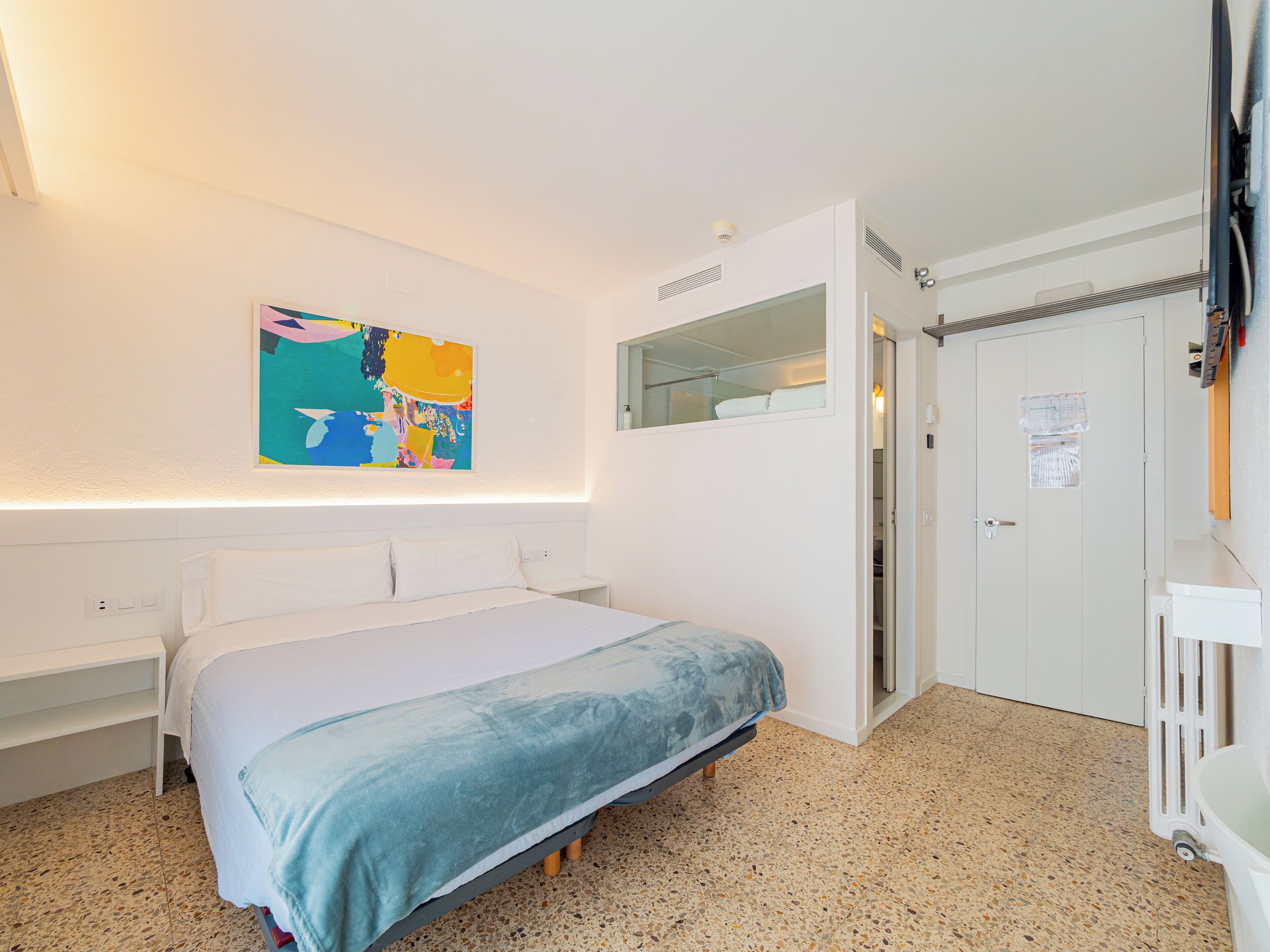 A bedroom with a bed, a white wardrobe, a bathroom with a mirror, and a colorful abstract painting on the wall.