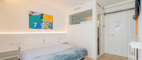 A bedroom with a bed, a white wardrobe, a bathroom with a mirror, and a colorful abstract painting on the wall.