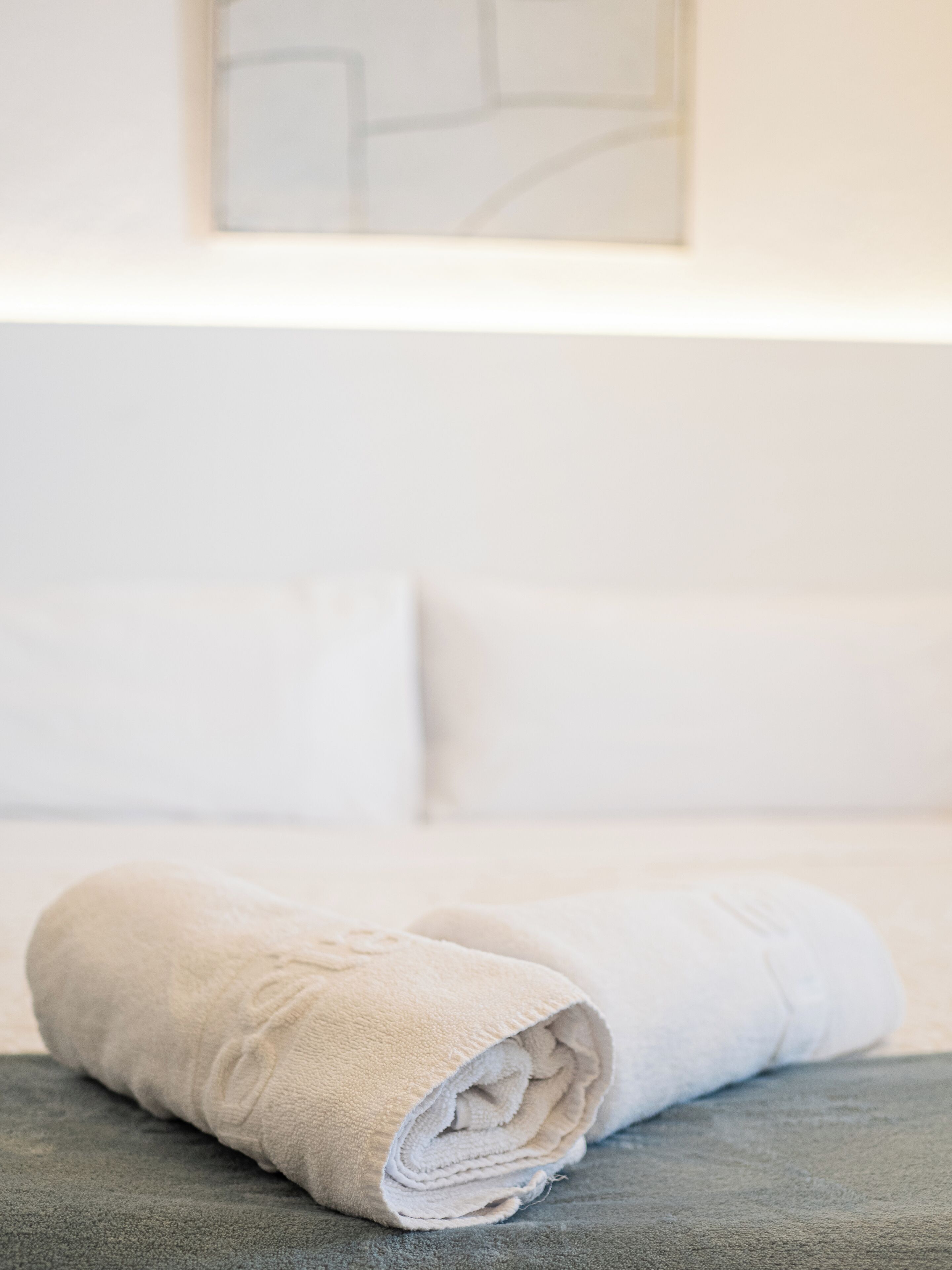 A neatly folded beige towel on a grey surface, likely a bed or couch, in a room with white bedding and a geometric wall art piece.