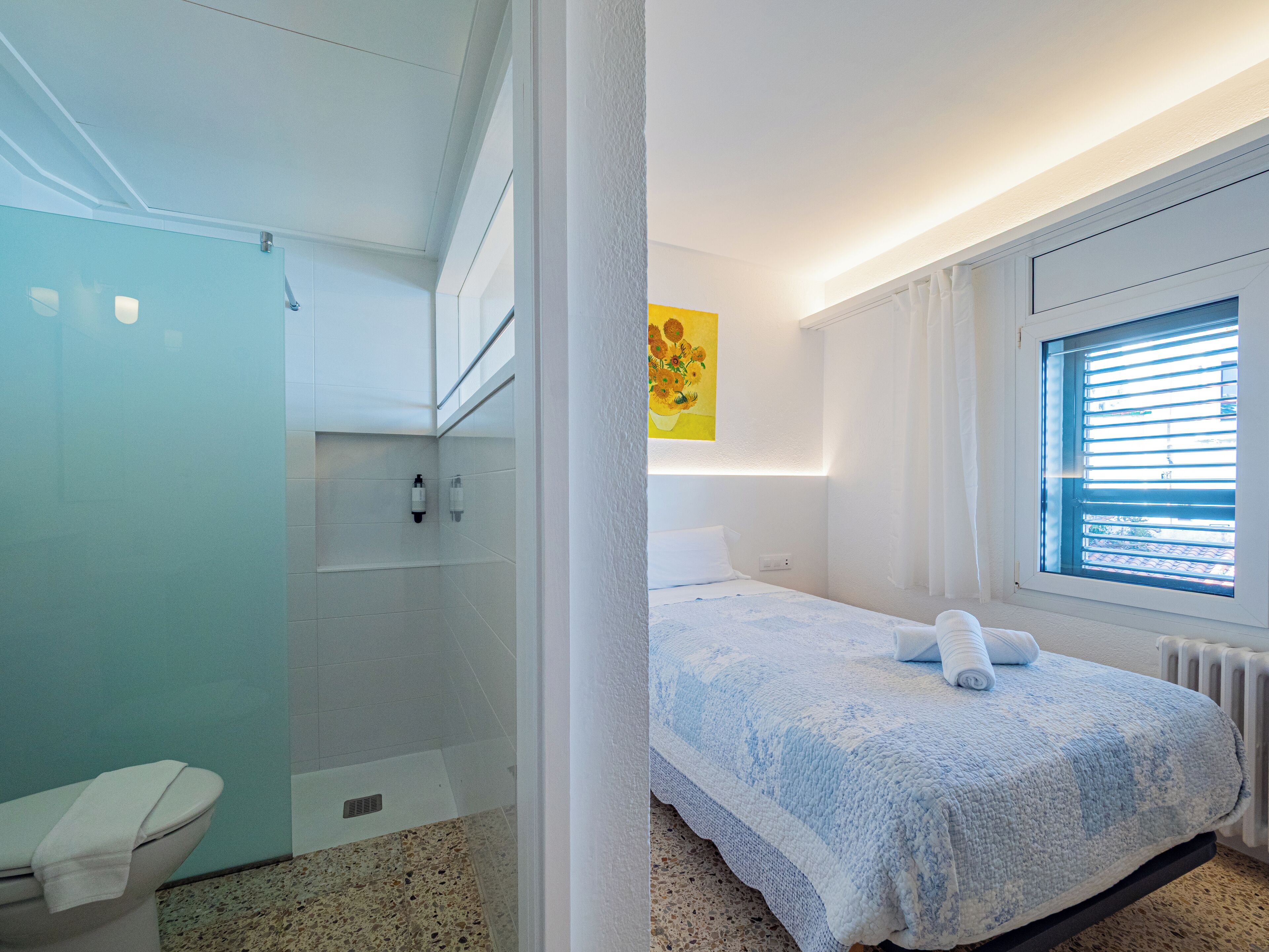 A modern hotel room with a shower, a bed with a blue blanket, a window with blinds, and a painting on the wall.