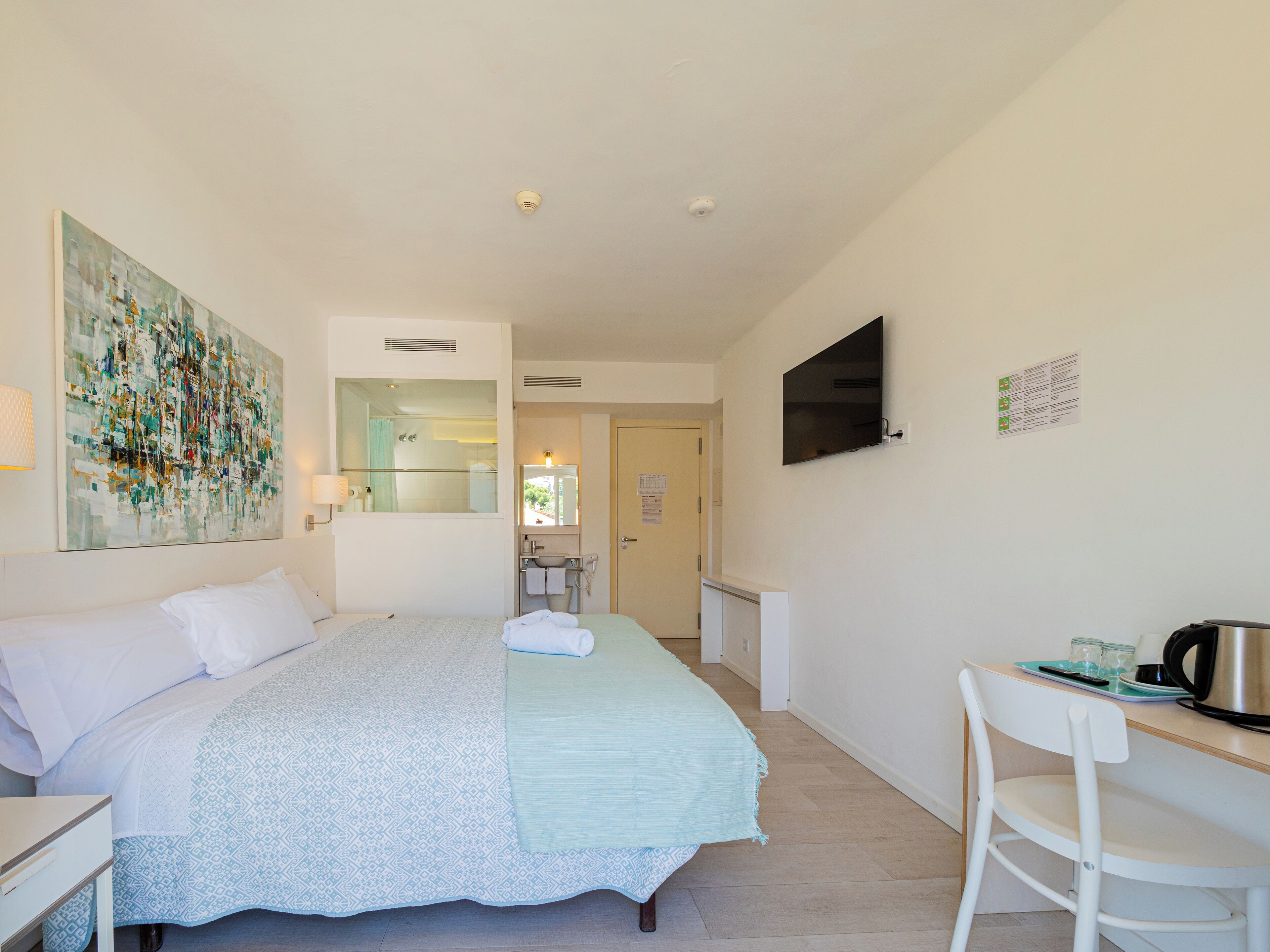 A hotel room with a bed, a desk, a chair, a TV, and a bathroom visible through an open door.