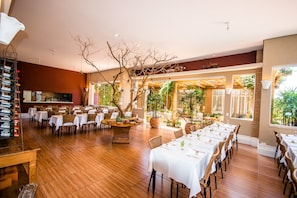 Uma ampla sala de jantar com várias mesas preparadas para hóspedes, com piso de madeira e iluminação natural proveniente de grandes janelas.