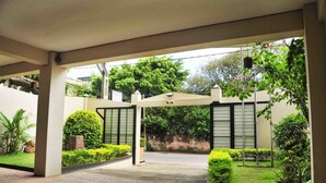 A covered outdoor area with a paved walkway, a small garden with neatly trimmed bushes and a spherical hedge, and a metal gate. - The Prestige Court Residence (Mount Lavinia)