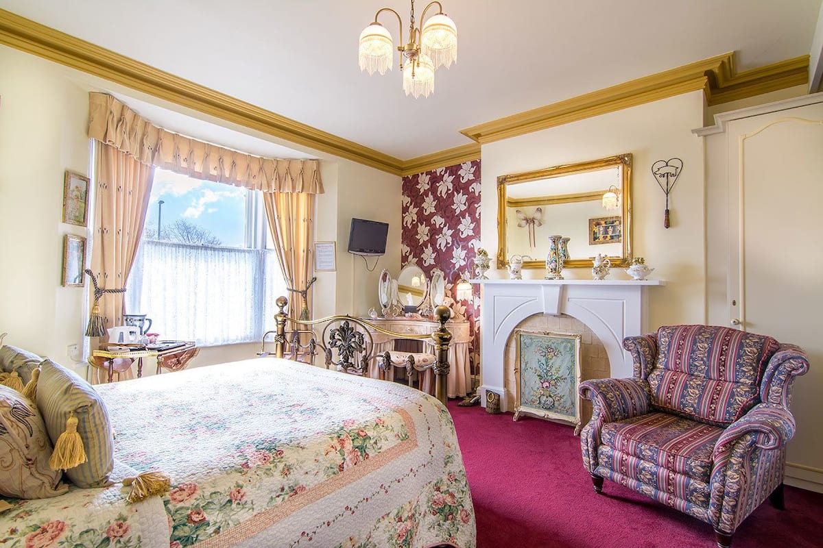 Une chambre d’hôtel classique, dotée d’un lit, d’une coiffeuse, d’une cheminée et d’un fauteuil à motifs.